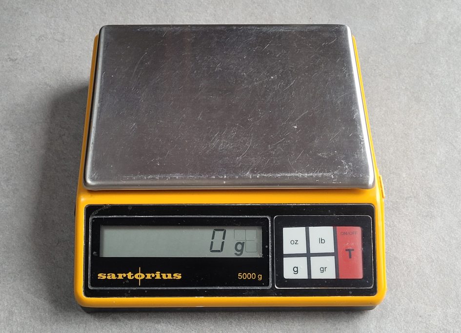 Top view looking down on a Sartorius electronic toploader 1006 MP9 balance with painted yellow metal case and large stainless steel balance plate. LCD display reads 0g. Push buttons select oz, lb, g, gr and tare.