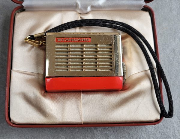Shows the gold plated grille and red plastic case with a carry stop on the left hand side. The radio sits in a satin lined clamshell case.